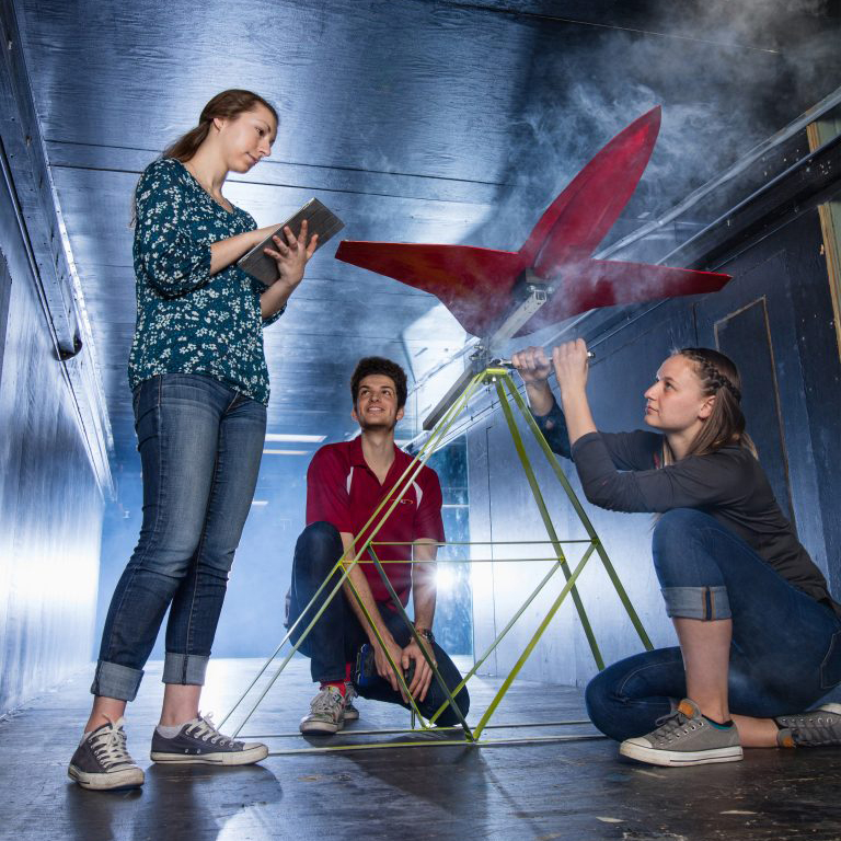 Three students working on model plane in wind tunnel, one with ipad, one with tool, one onlooking