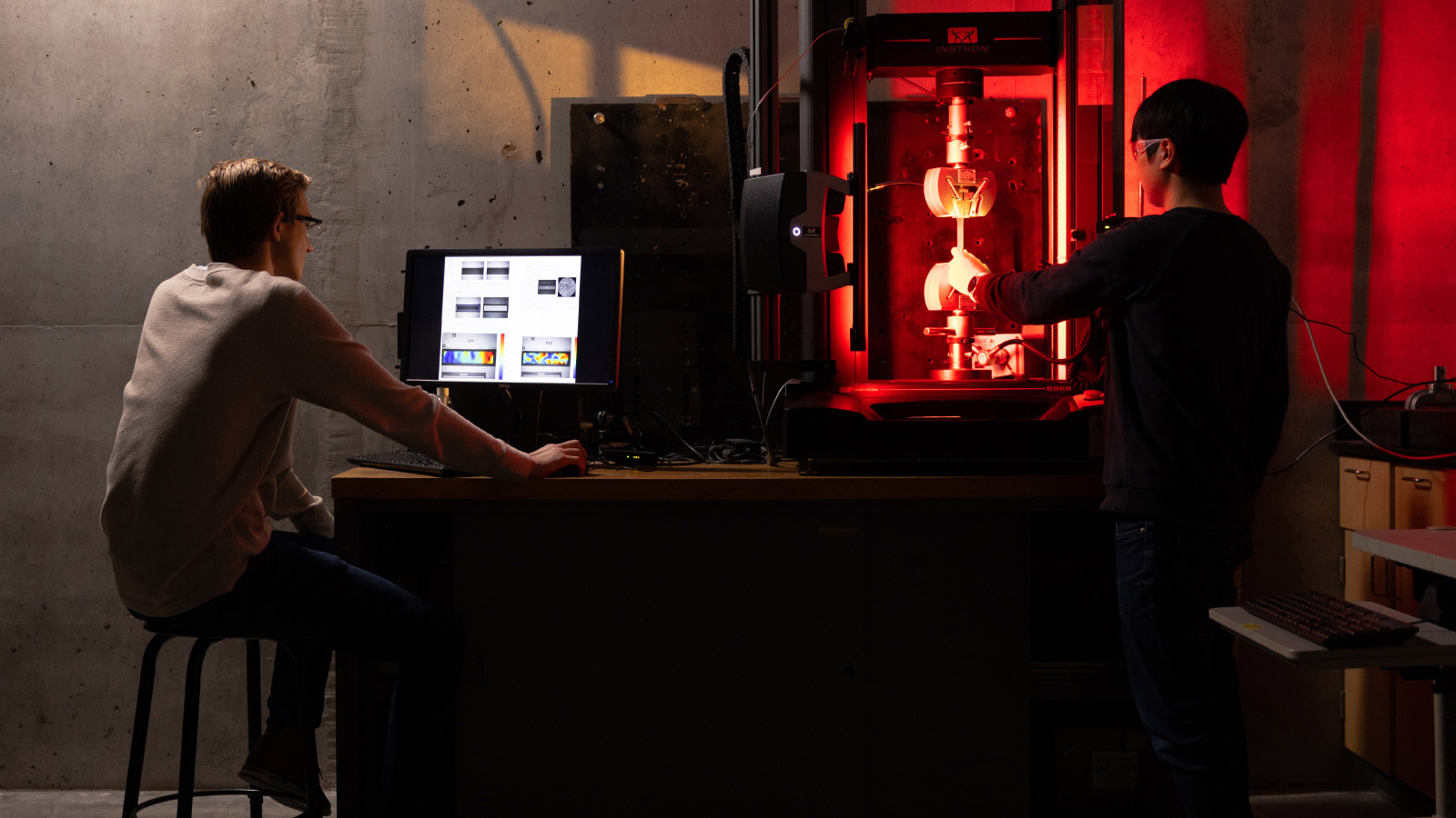 Two students working a computer and machine, glowing red